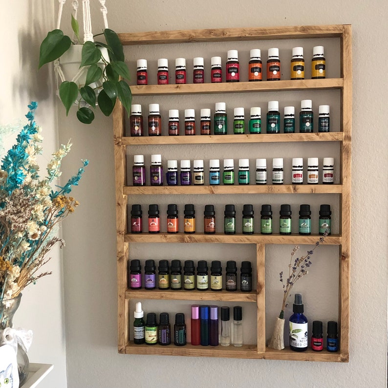 May include: A wooden shelf unit with five shelves for storing essential oil bottles. The shelves are filled with rows of small glass bottles with white lids. The bottles are arranged in a rainbow pattern of colors, from red to purple. The shelf unit is made of natural wood and has a rustic finish.