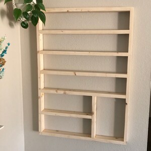 May include: A wooden wall shelf with six shelves and a small compartment on the bottom right. The shelf is made of unfinished wood and is mounted on a white wall.