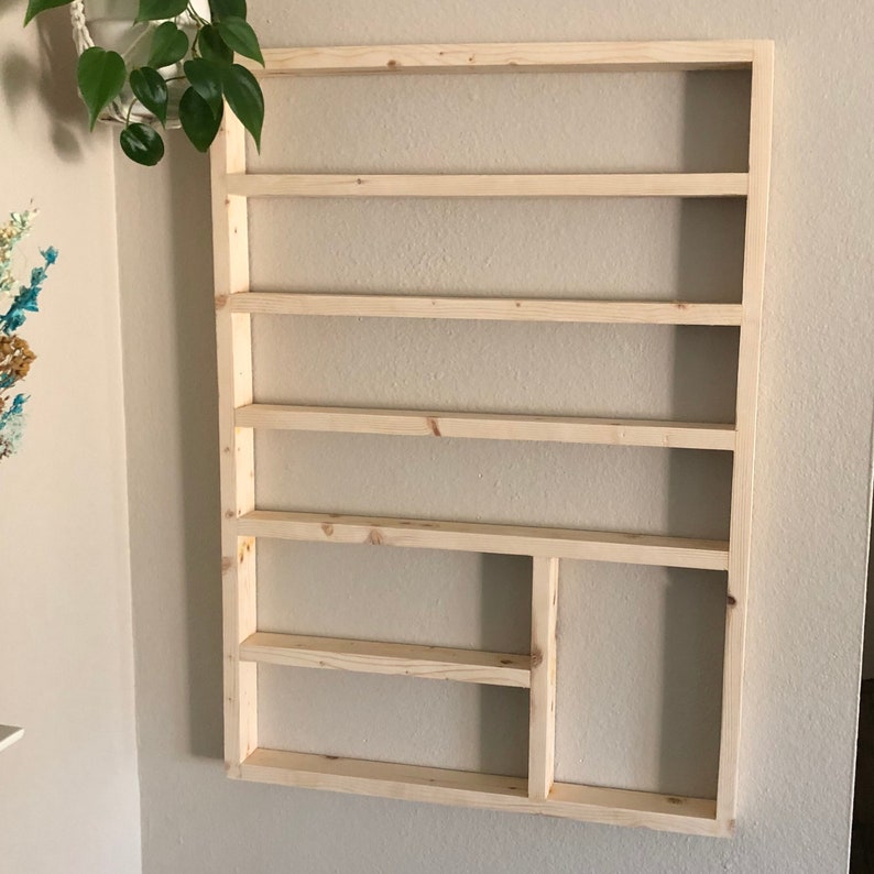 May include: A wooden wall shelf with six shelves and a small compartment on the bottom right. The shelf is made of unfinished wood and is mounted on a white wall.