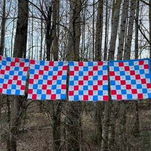 May include: Four square quilts with a red, white, and blue checkerboard pattern are hanging from tree branches. The quilts are displayed outdoors, with a forest background. The quilts are made of fabric.