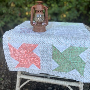 May include: A vintage quilt with pinwheel designs in coral and mint green, set on a white chair. The quilt has a floral pattern and is topped with a rusty lantern, creating a rustic, country aesthetic.