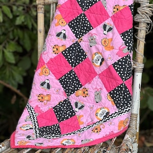 May include: A pink and black baby quilt with a patchwork design featuring various animal illustrations. The quilt is folded and draped over a wicker chair.
