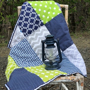 May include: A patchwork quilt with a variety of patterns in shades of blue, green, and gray. The quilt is folded and placed on a white wicker chair. A small, blue oil lamp sits on top of the quilt.