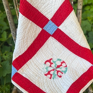 May include: A handmade quilt with a white background, red stripes, and blue squares. The quilt features a circular design with red and white polka dots and a light blue and white floral pattern. The quilt is draped over a wooden chair.