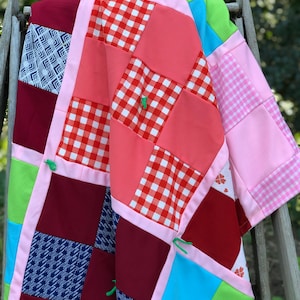 May include: A patchwork quilt with squares of red, pink, blue, green, and white fabric. The quilt is folded and draped over a wooden chair.