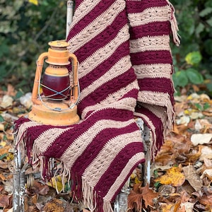 May include: A crocheted striped throw blanket in burgundy and beige with fringe on the edges. The blanket is draped over a wooden chair with a vintage lantern sitting on the seat.