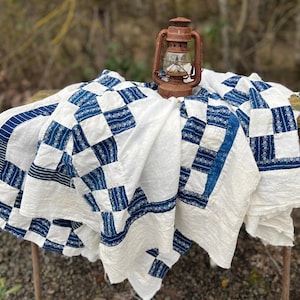 May include: A vintage quilt with a white background and blue checkered pattern is draped over a wooden surface. A rusty lantern sits atop the quilt. The quilt's design features squares and stripes.