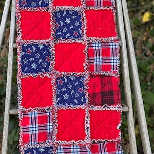 May include: A rectangular rag quilt runner featuring a patchwork design of red, navy blue with white star patterns, and red, white, and blue plaid fabric squares. Each square is stitched with an X pattern and has frayed, exposed seams.