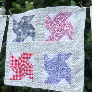 May include: A white quilt with four squares, each featuring a different patterned fabric in a windmill design. The fabrics are blue and white plaid, pink and white floral, red and white gingham, and blue and white floral.
