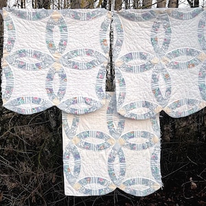 May include: Three white quilts with a double wedding ring pattern, featuring floral and striped fabric accents in blue, green, and pink. The quilts are hanging outdoors, showcasing a vintage aesthetic.