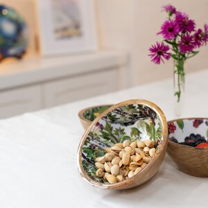 May include: Three wooden bowls with floral patterns. The bowl on the left contains pistachios, the middle bowl is empty, and the bowl on the right contains blackberries and strawberries.