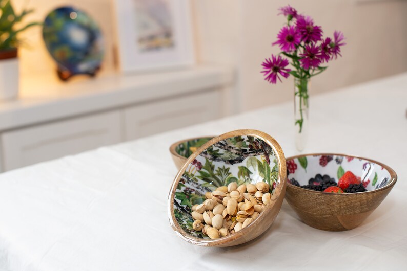 May include: Three wooden bowls with floral patterns. The bowl on the left contains pistachios, the middle bowl is empty, and the bowl on the right contains blackberries and strawberries.