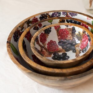 May include: Set of three wooden bowls with a white background and a colorful berry design. The bowls are nested inside each other, with the largest bowl on the bottom and the smallest bowl on top.