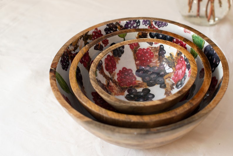 May include: Set of three wooden bowls with a white background and a colorful berry design. The bowls are nested inside each other, with the largest bowl on the bottom and the smallest bowl on top.