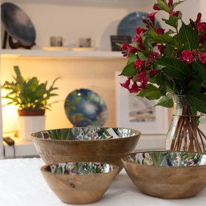 May include: Three wooden bowls with a floral design on the inside. The bowls are nested inside each other, with the largest bowl on the bottom and the smallest bowl on top. The bowls are on a white tablecloth.
