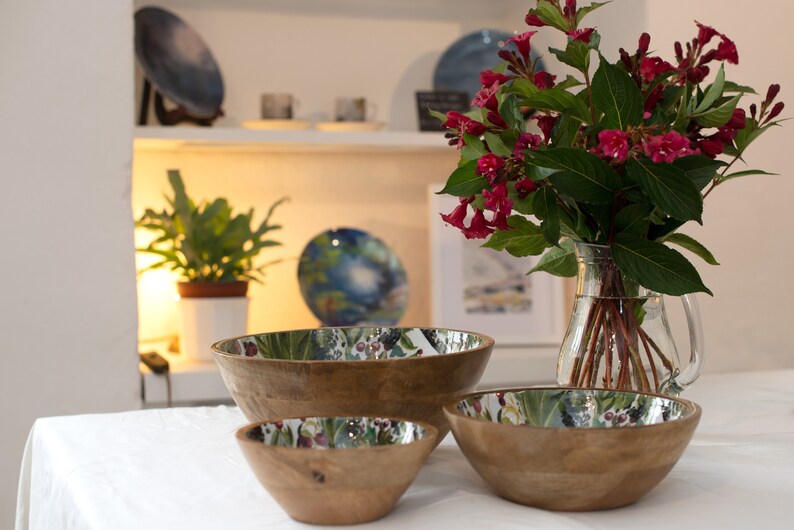 May include: Three wooden bowls with a floral design on the inside. The bowls are nested inside each other, with the largest bowl on the bottom and the smallest bowl on top. The bowls are on a white tablecloth.