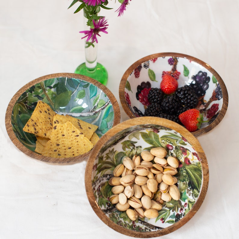 May include: Three wooden bowls with floral designs. The bowls are filled with snacks, including tortilla chips, pistachios, and berries.