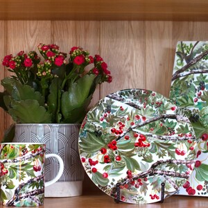 May include: Set of four white ceramic mugs with a green and red floral pattern. The mugs are arranged in a row on a wooden shelf. A larger white ceramic plate with the same floral pattern is on the shelf to the right of the mugs. A square white ceramic platter with the same floral pattern is on the shelf to the right of the plate. A potted plant with red flowers is on the shelf to the left of the mugs.