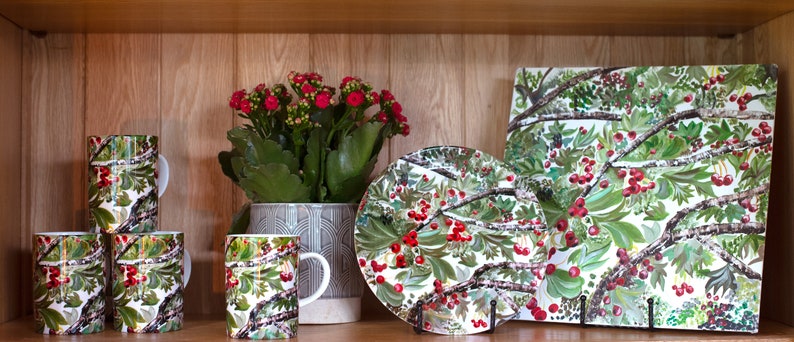 May include: Set of four white ceramic mugs with a green and red floral pattern. The mugs are arranged in a row on a wooden shelf. A larger white ceramic plate with the same floral pattern is on the shelf to the right of the mugs. A square white ceramic platter with the same floral pattern is on the shelf to the right of the plate. A potted plant with red flowers is on the shelf to the left of the mugs.