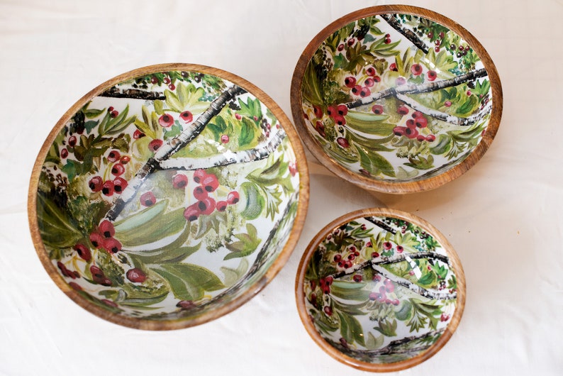 May include: Three wooden bowls with a white background and a floral pattern of green leaves, red berries, and white branches. The bowls are nested inside each other, with the largest bowl at the bottom and the smallest bowl at the top.