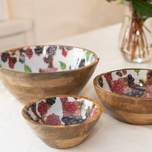 May include: Three wooden bowls with a white background and a colorful berry pattern. The bowls are nested inside each other, with the largest bowl at the bottom and the smallest bowl at the top.