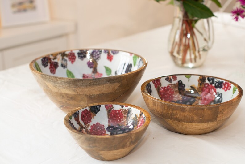 May include: Three wooden bowls with a white background and a colorful berry pattern. The bowls are nested inside each other, with the largest bowl at the bottom and the smallest bowl at the top.