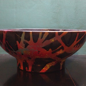 May include: A large, black ceramic bowl with a wide, rounded base. The bowl is decorated with a splatter pattern of red, orange, yellow, and blue paint.