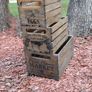 May include: Three stacked wooden crates with dark brown stain. The crates have cut-out handles and printed text: "Farm Fresh Eggs," "Farm Fresh Milk," and "Fresh Flower Market." The crates are sitting on a bed of wood chips.
