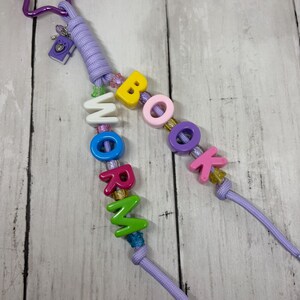 May include: A purple star-shaped carabiner attached to a lavender cord featuring a small purple book charm and multi-colored letter beads spelling out BOOK WORM. The design includes various small accent beads in shades of pink, yellow, and green.