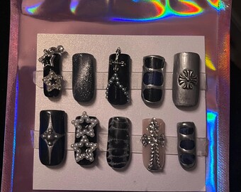 May include: A set of ten black and silver press-on nails with various designs, including stars, crosses, and geometric patterns.