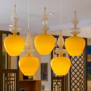 Może przedstawiać: Pięć lamp wiszących z żółtymi, zaokrąglonymi szklanymi kloszami i przezroczystymi szklanymi akcentami. Światła zwisają z sufitu na cienkich białych sznurkach. Tło obejmuje drewniane przegrody pomieszczeń i jadalnię.