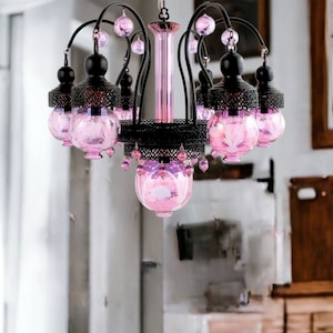 May include: A decorative chandelier with a black metal frame and pink glass globes. The chandelier has a central pink column and is adorned with pink crystal accents. The design is ornate and elegant, suitable for interior lighting.