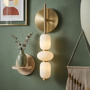 Puede incluir: Una lámpara de pared moderna con acabado de latón y tres luces de vidrio esmerilado. La lámpara está montada en una pared verde, junto a una pequeña estantería con un jarrón y una obra de arte enmarcada.