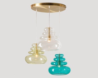 May include: A modern pendant light fixture with three stacked glass shades in clear, teal, and yellow-green. The shades are suspended from a brass-colored circular canopy. The light fixture is designed for indoor use.