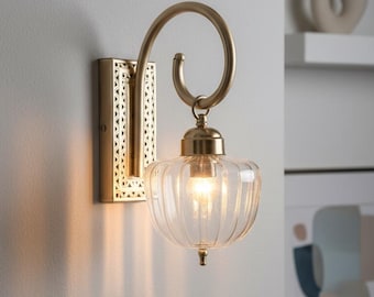 May include: A wall sconce featuring a brushed gold metal finish with a rectangular backplate displaying a geometric cutout pattern. A curved arm supports a clear, ribbed glass shade shaped like a pumpkin, housing a single glowing light bulb.