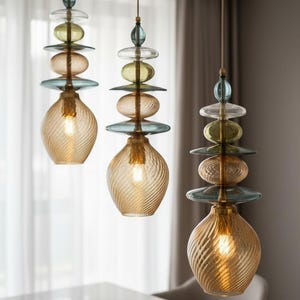 May include: Three pendant lights hanging from thin cords, each featuring a stack of colorful glass elements in blue, clear, and olive green, topped by a textured, amber-colored glass bulb shade with a spiral pattern and an internal light source.