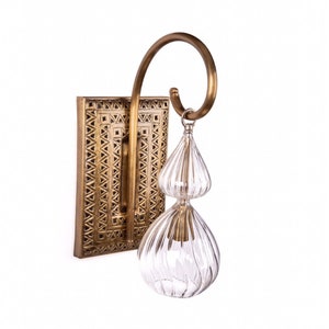 May include: A brass wall sconce with a rectangular backplate featuring a geometric pattern. A curved brass arm supports a clear, ribbed glass pendant with two bulbous sections. The sconce is designed for indoor lighting.