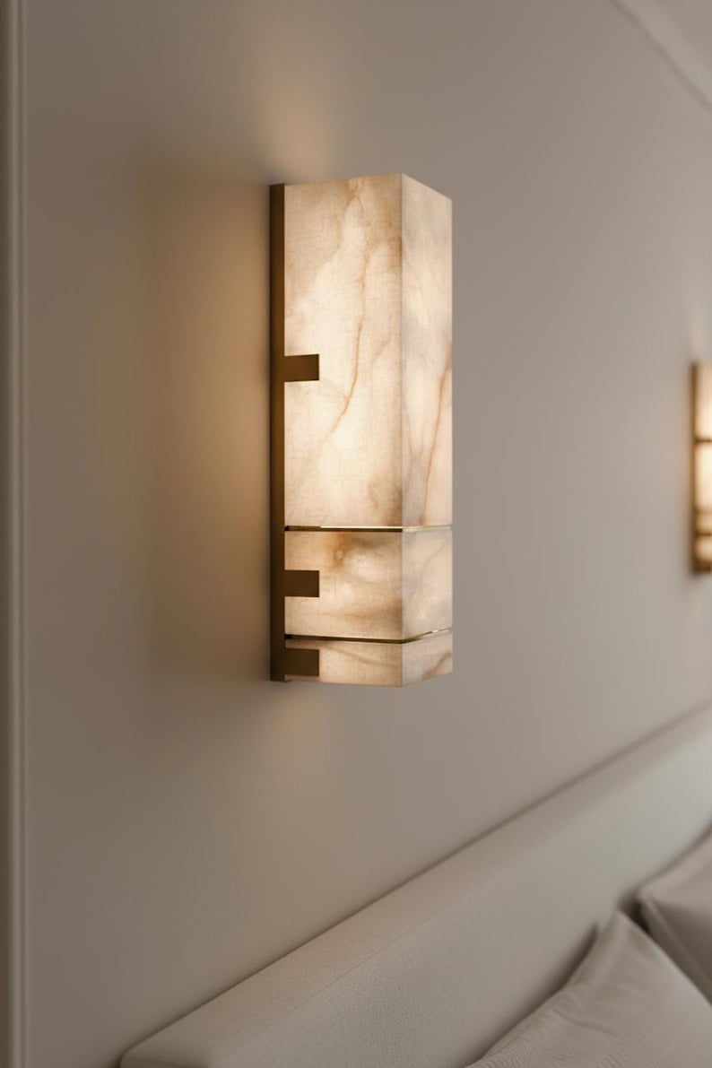 Op de afbeelding: Een rechthoekige wandlamp met een marmerachtige uitstraling. De lamp heeft een warm, gloeiend licht en is gemonteerd op een neutraal gekleurde muur. De armatuur heeft een bronzen metalen frame met een geometrisch ontwerp.