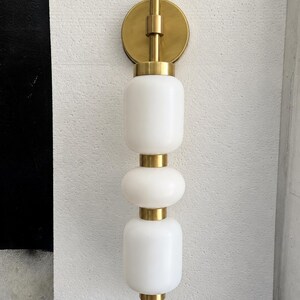 May include: A wall-mounted light fixture featuring a vertical arrangement of three white frosted glass globes separated by brushed brass metal spacers. The fixture is attached to the wall with a circular brushed brass mounting plate.