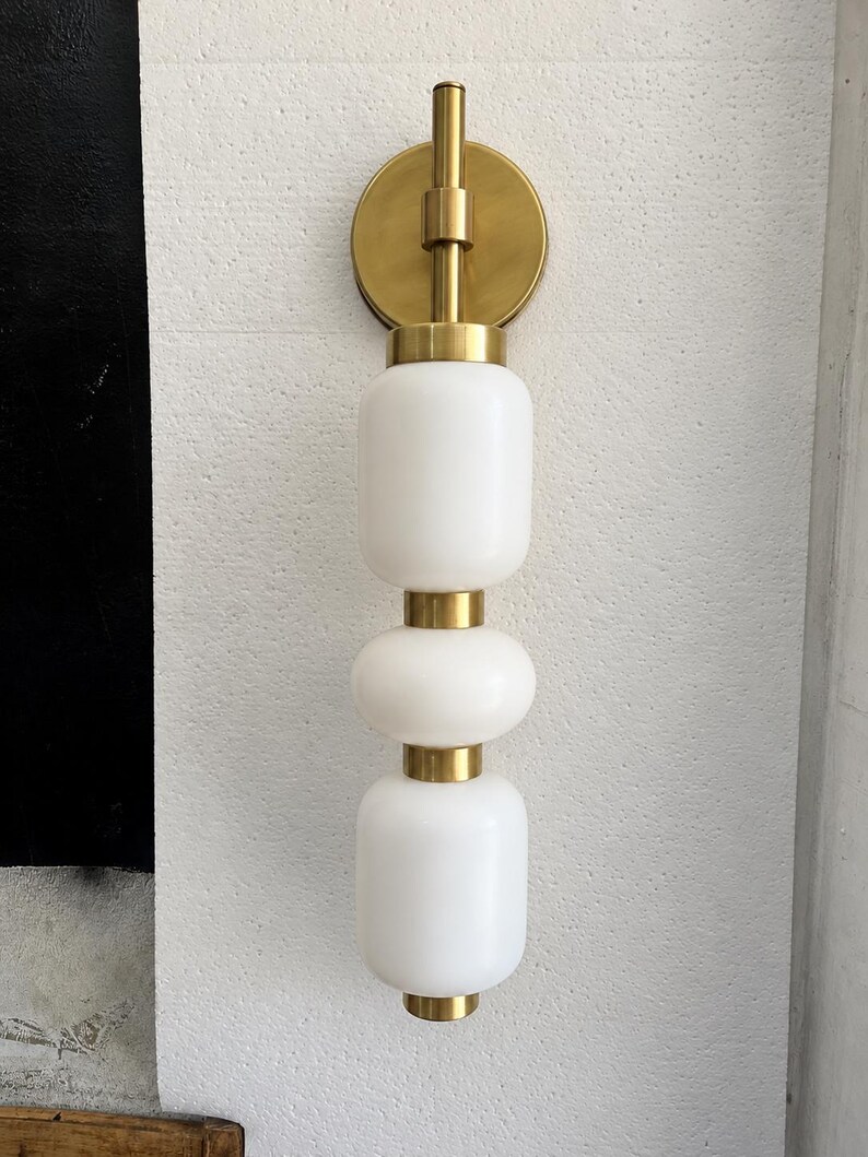 May include: A wall-mounted light fixture featuring a vertical arrangement of three white frosted glass globes separated by brushed brass metal spacers. The fixture is attached to the wall with a circular brushed brass mounting plate.