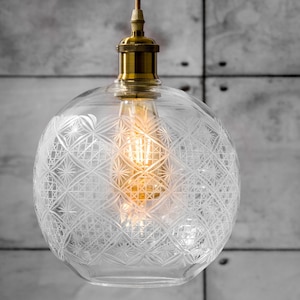 以下が含まれることがあります： 装飾的なエッチング模様が施された透明なガラスペンダントライト。真鍮色のトップとヴィンテージスタイルのエジソン電球を備えています。ゴールドカラーのコードから吊り下げられています。