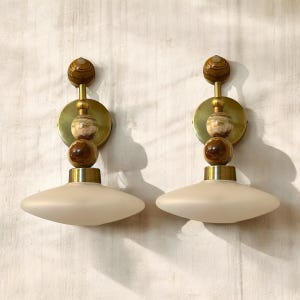 May include: Two wall sconces with a mid-century modern design. Each sconce features a brass circular backplate, a series of decorative spheres in brown and beige tones, and a frosted, saucer-shaped glass shade. The sconces are mounted on a textured wall.