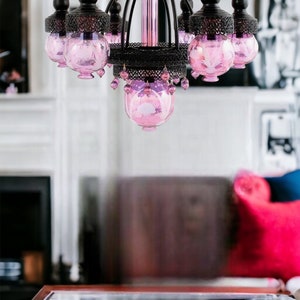May include: A decorative chandelier with a black metal frame and pink glass globes. The chandelier has multiple arms with hanging pink crystal accents. The central column is pink, and the fixture is designed for indoor lighting.