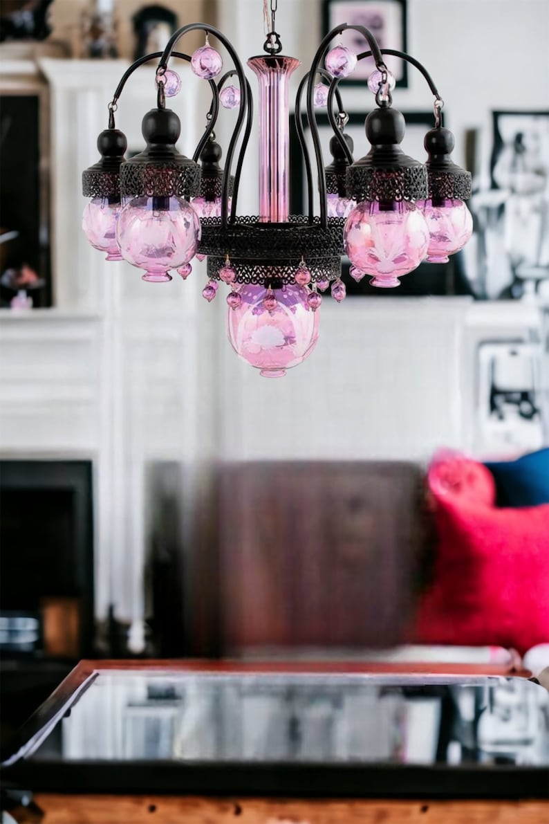 May include: A decorative chandelier with a black metal frame and pink glass globes. The chandelier has multiple arms with hanging pink crystal accents. The central column is pink, and the fixture is designed for indoor lighting.