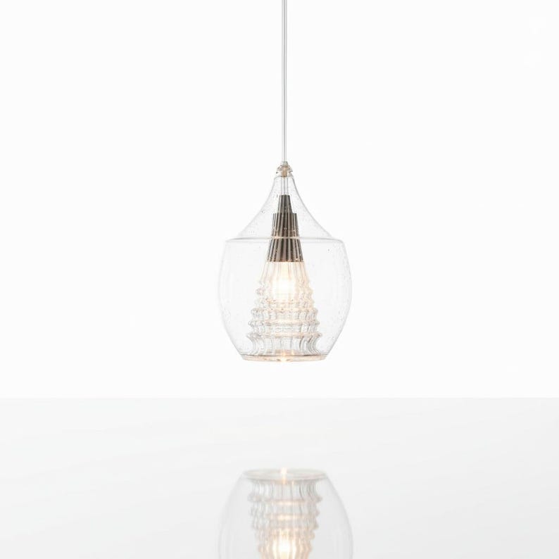 May include: Clear glass pendant light with a teardrop-shaped shade. The light fixture has a textured glass exterior and a decorative interior element. The light hangs from a thin white cord, creating a modern and elegant aesthetic.
