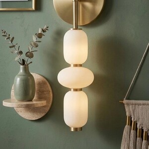May include: A wall-mounted light fixture featuring a vertical arrangement of three frosted white glass globes separated by brushed brass accents, attached to a circular brass backplate with a slender central rod.