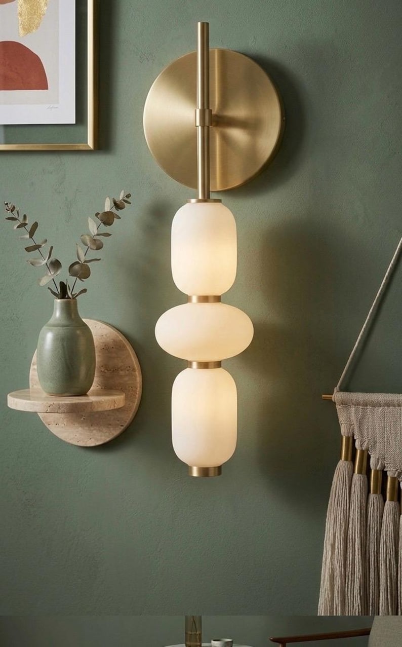 May include: A wall-mounted light fixture featuring a vertical arrangement of three frosted white glass globes separated by brushed brass accents, attached to a circular brass backplate with a slender central rod.