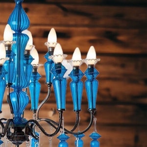 May include: A close-up of a blue glass chandelier with multiple light bulbs. The chandelier has a dark metal frame and is adorned with blue glass accents. The background is a wooden wall.