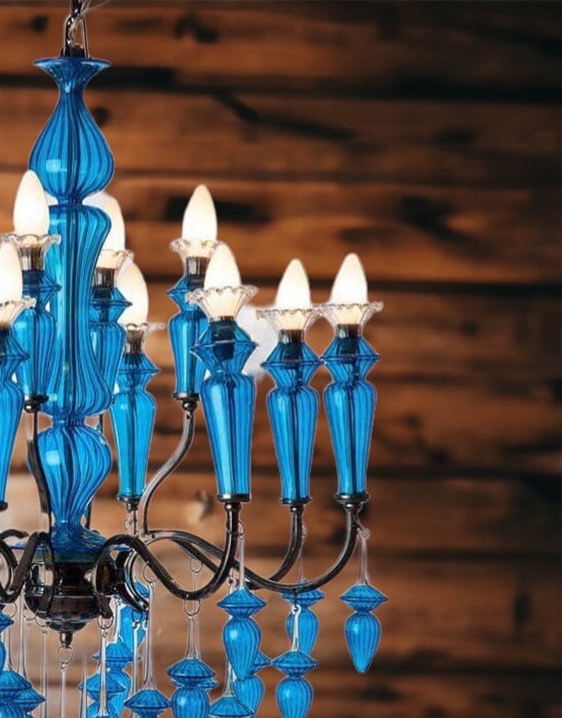 May include: A close-up of a blue glass chandelier with multiple light bulbs. The chandelier has a dark metal frame and is adorned with blue glass accents. The background is a wooden wall.