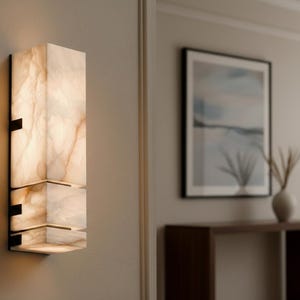 Op de afbeelding: Een moderne wandlamp met een rechthoekig, gelaagd ontwerp. De lamp is gemaakt van een doorschijnend, cr&egrave;mekleurig materiaal met bruine adering, gemonteerd op een zwart metalen frame. De lamp is verlicht en geeft een warme gloed.